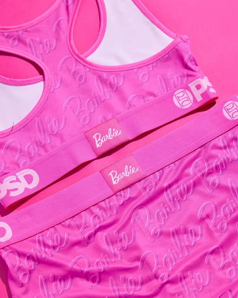 Bright pink sports bras with Barbie logo print and text labels, displayed flat lay on matching background.