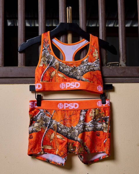 Realtree Edge orange camouflage sports bra and boy shorts set with tree branch pattern and PSD branding on hanger.