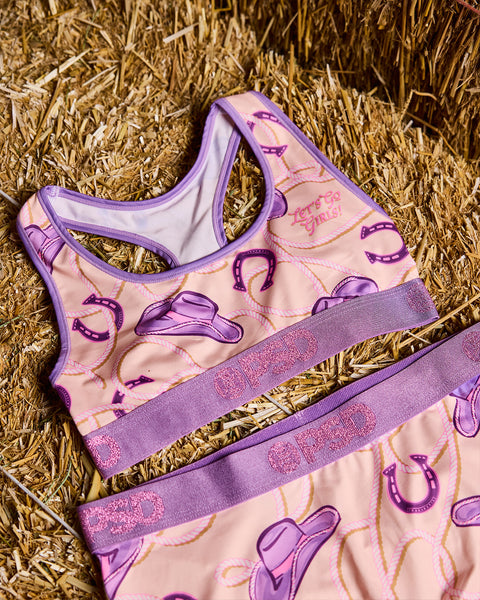Purple and pink sports bra with cowboy hat and horseshoe print, purple trim and glitter waistband on straw background.