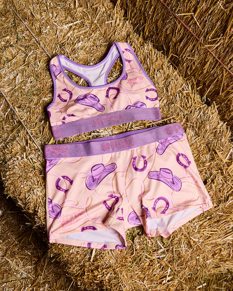 Two-piece children's swimsuit with purple cowboy hats and horseshoes on pink fabric, displayed on straw background.