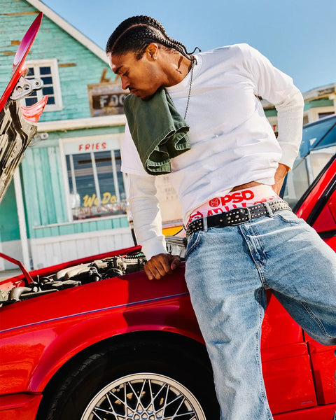 Person wearing white long-sleeve shirt and studded black belt leaning against open red car hood at beachside location.